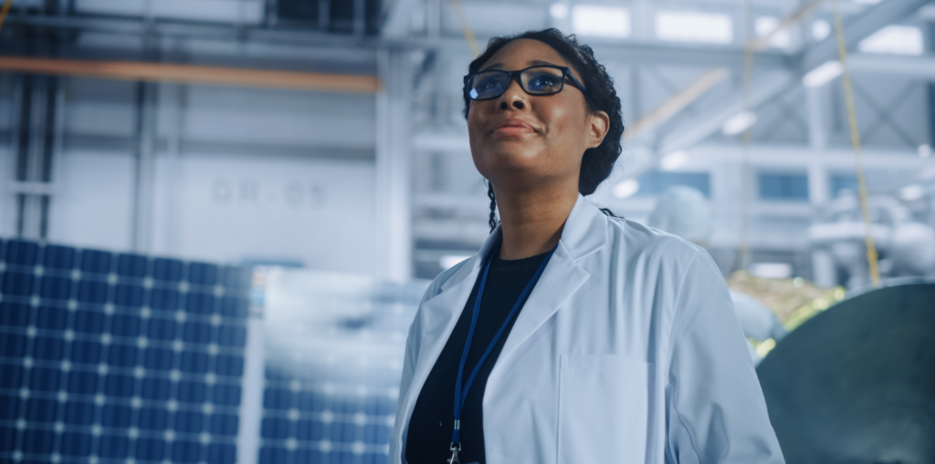 Woman with glasses and nose piercing wearing lab coat and lanyard, standing inside manufacturing facility